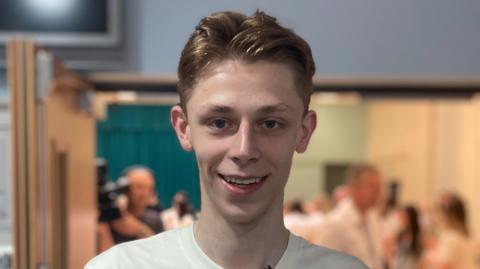 Harry McClelland wears a white T-shirt and has medium length light brown hair. He is looking straight at the camera and the background is blurred.