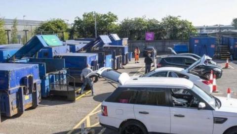 A group of cars parked at a household waste recycling centre with a number of large waste containers in blue to the left of the image.