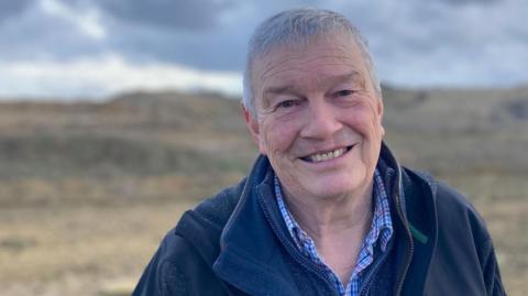 Stephen Adkins in a black fleece, blue jumper and checked shirt in front of a fence with a field behind