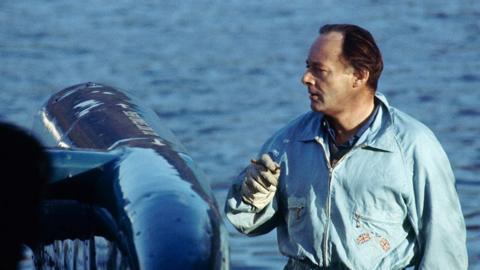 DOnald Campbell, wearing overalls and smoking a cigar, stands next to Bluebird. He has dark hair, brushed back from his face.