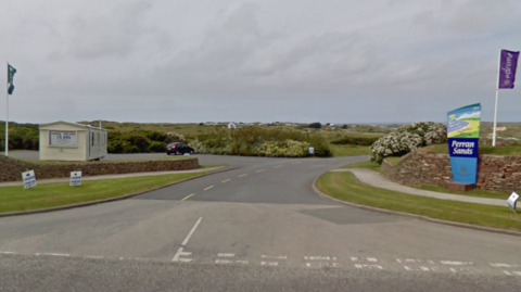 The entrance to the holiday park. There is a sign with an artistic representation of the bay and beach and the words PERRAN SANDS below it on one side and a static caravan on the other side in a car park which has one car in it. There are flags either side of the entrance which is flanked with grass verges and paths. Beyond the drive are bushes and in the distance buildings.