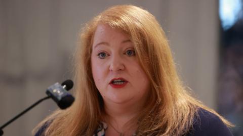 A long red-haired woman speaks into a black microphone, wearing red lipstick. She wears a purple cardigan with a white and teal shirt underneath. 