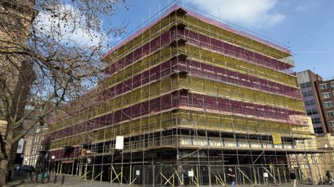 The outside of Higham House - a large building covered completely in scaffolding with yellow and pink netting over the outside. There is a man walking in front of it on his phone and a leafless tree to the left.