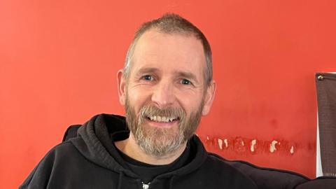 A man with short brown and grey hair and a beard wearing a black hoodie sits against a red wall. He is smiling at the camera