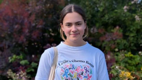 Smiling teenage girl Lexie. Her brown hair is tied back from her face and she has a centre parting. She is wearing a white T-shirt printed with the words Lexie's Chemo Bags. The straps of a cloth bag can been see over her right shoulder.