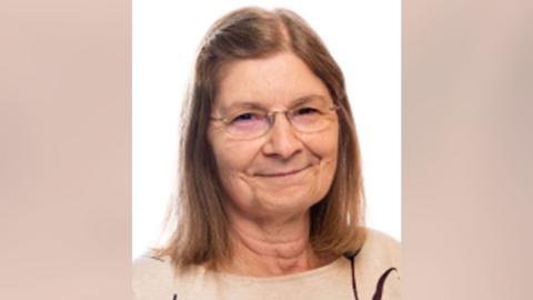 A woman with long light brown hair and wearing glasses looking into the camera. She is wearing a light brown jumper.