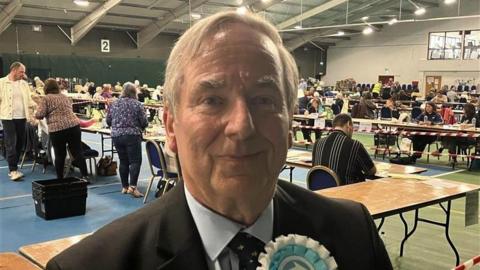 Reform's Robert Parsonage. He is wearing a black suit, blue shirt and has a Reform rosette on his chest. He has short grey hair.