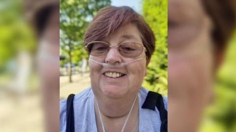 Sarah with a breathing tube in her nose smiling at the camera. She has short dark hair and is wearing a grey top. She is outside on a sunny day.