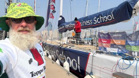 Bob is stood ashore with a big grey beard, sunglasses, and a green Leicester Tigers bucket hat on. He is stood in front of his racing yacht, which has a member of crew on board. It is sunny and there are no clouds in the photo. 
