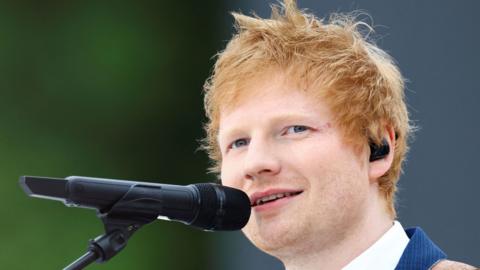 Ed Sheeran is pictured singing into a microphone looking away from the camera. He has red hair and wears a blue suit with a white shirt underneath. He wears a black earpiece. 