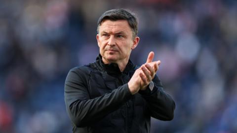 Paul Heckingbottom looks to his right and claps his hands together in front of his chest. He wears a black jacket and watch on his left wrist.