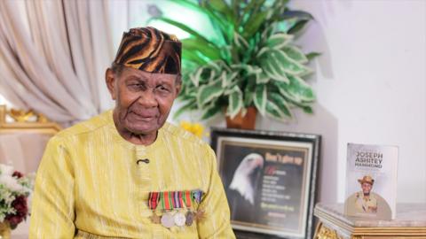 100-year-old Ghanian veteran Joseph Hammond sits reflectively beside his autobiography book on a table with potted plants behind him, while speaking to a reporter in an interview about his time fighting in Burma. He is wearing a black and brown striped hat and a yellow and white striped top with five war medals shaped as circles and stars pinned to it.
