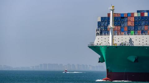 A green shipping container vessel appears on the right of the image, in front of a large cityscape in the background.
