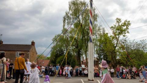 A tall maypole which is stripy blue, white and red has coloured ribbon coming out of it which children are pulling and dressed in traditional clothing. People can be seen stood around the maypole watching the children.
