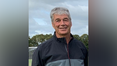 A man with short grey hair standing in front of a football net. He is wearing a black and grey tracksuit and smiling.