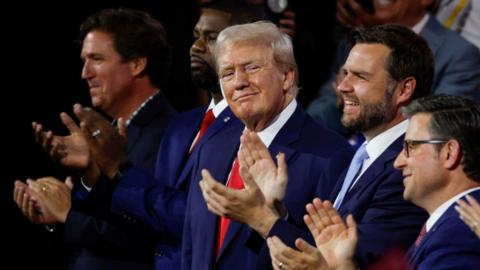 Donald Trump turns to his left with a smile as Tucker Carlson, Byron Donalds, JD Vance and Mike Johnson stand on either side of him, all wearing similar blue suits, and applauding as they face an unseen scene before them