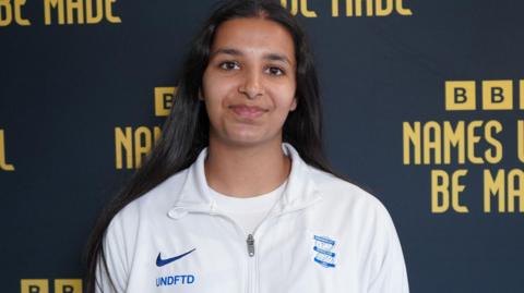 Riya Mannu, a woman wearing a white t-shirt and white sports jacket is stood against a BBC Names Will Be Made step and repeated board which is navy blue with yellow lettering.