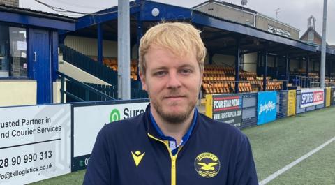 Gary Shufflebottom, who has blonde hair, is wearing a navy and yellow Bangor FC tracksuit top. He is standing on the pitch at Clandeboye Park, near a stand with orange and blue seats.