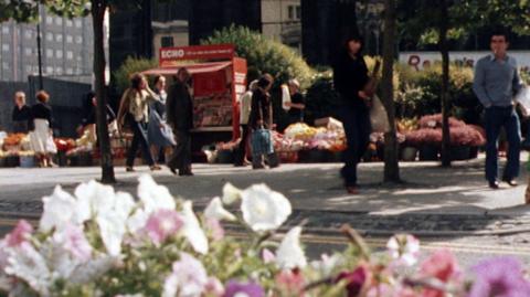 Low angle shot of people walking along the pavement in Liverpool past a bed of flowers and a Liverpool Echo branded magazine stand.