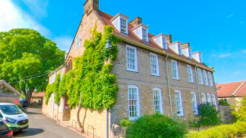 A large, three-storey brick building with tall, white windows, with a tree in the background and bushes at the front 