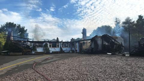 A bungalow that is charred and blackened with parts of its roof collapsed or missing due to a fire. Grey smoke rises from the remains.