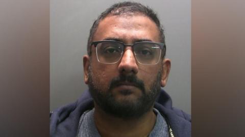 A mugshot of Shamir Chandran. He has short salt and pepper hair and a trimmed beard. He wears glasses with a blue hoodie and grey t-shirt.
