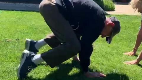 An elderly man has his hands on grass with bent knees facing the ground in a yoga position