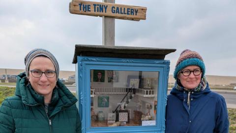 (Left to right) Antje Rook smiling at the camera and wearing a green puffa jacket and beanie hat and Yollande Posthumus is also smiling at the camera wearing a blue raincoat and a knitted hat. In between them is a beehive box where the tiny gallery can be viewed.