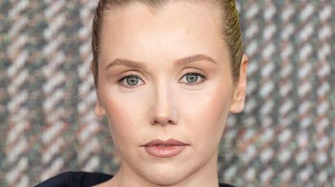 A glamorous red carpet shot of actress Lauren Lyle is a close-up of her face, with a tweed-effect on the background behind her.