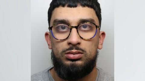 A mug shot of Hassan Hussain, a man with black hair and a black beard, wearing glasses and looking straight at the camera.