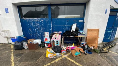 Household items, such as a doll's house, plastic bags and a desk next to  a blue garage door on the wet ground after rain