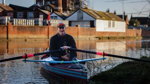 Mike Ellicock sits in his boat, a combination of a sea kayak and an ocean rowing boat, and holds the oars. He is looking at the camera, wearing a dark blue jumper and baseball cap, with sunglasses rested on the peak of the cap. He is on a river with buildings behind him.