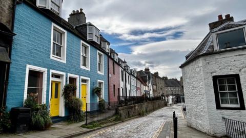 a bright blue house with yellow doors, next to a range of other brightly coloured houses. The house is situated on a cobble-stoned street and is contrasted by a black and white house directly adjacent.