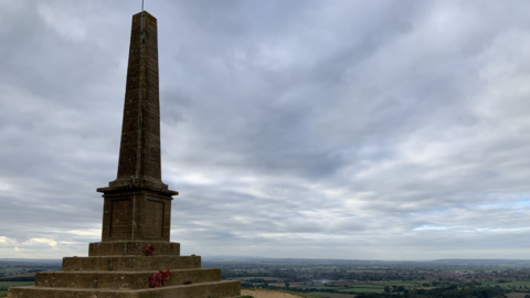 A view from behind the War Memorial, looking out over the landscape below Ham Hill. The memorial itself consists of four steps with a large stone obelisk in the middle. The names of the dead are written on tablets on its side.