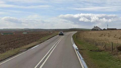 A single carriageway road is surrounded by flat fields with hills in the distance. There are double white lines in the centre of the road to show that overtaking is prohibited.