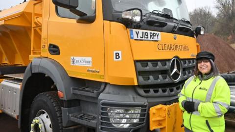 A woman who is a gritter driver dressed in orange and yellow hi-vis stands next to a gritter 