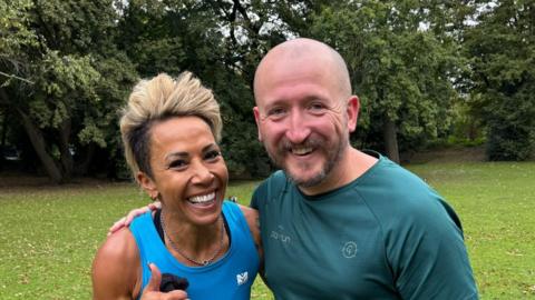 Two people wearing sport clothing pose for a photo with their arms around one another. The woman on the left is wearing a blue vest and has short blonde hair swept over her head. The man on the right is wearing a dark green top and has a bald head and dark coloured beard.