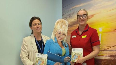 Abby, a woman with brown hair and a cream jacket, a lifesize cutout of Dolly Parton (a woman with blonde hair and a blue blazer), and Regan, a woman in a red uniform. All are carrying books.