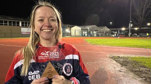 Debbie Armstrong is wearing her running gear and holding the award she received as Volunteer of the Year. She has long blonde hair and is smiling, looking happy. The athletics track is lit up behind her.