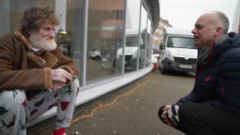 A man in a brow coat and pyjama bottoms smokes a cigarette as he asked questions by BBC South health correspondent Alastair Fee