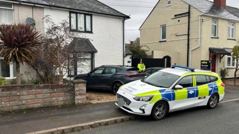A police car with two tyres on the pavement outside a semidetached home. It is a white house with black trim windows. There is a grey sports car parked on the homes gravel drive where a police officer woith a bright yellow jacket is standing with his arms crossed