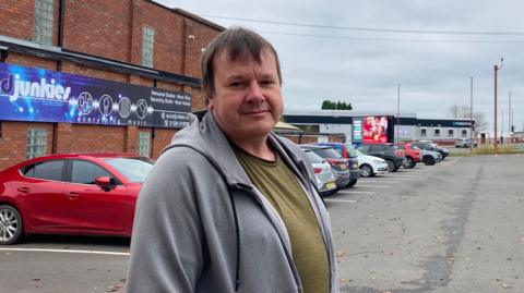 A man wearing a green t-shirt and a grey hooded top is stood in front of a car park and a red brick building with business branding and signage on it.