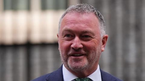 Steve Reed speaking. He wears a white shirt, a spotty green tie and a navy blazer