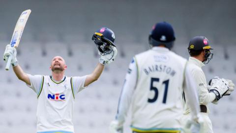 Yorkshire batsman Adam Lyth holding his bat in his right hand and helmet in his left hand in celebration while looking skywards after scoring a century against Surrey with Jonny Bairstow in the foreground ready to greet him.