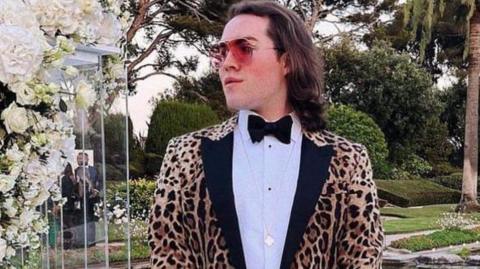 Jack Watkin is holding a handbag while wearing a leopard print jacket, white shirt and black bow tie. He is also wearing sunglasses in this social media image taken in a landscaped garden with large pond.