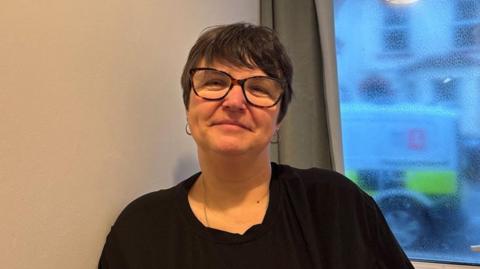 Michelle Grimes smiles looking directly into the camera. She has short brown hair and is wearing a black t-shirt, silver necklace, a pair of hooped earrings and tortoiseshell glasses.