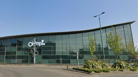 The exterior of the Olympia Leisure Centre, a large glass-sided building with a road in the foreground 