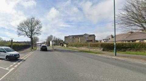 A wide paved road in a suburban or residential area during daylight.

In the foreground, the road occupies most of the image. White lane markings and a painted roadside area can be seen on the left-hand side. A small white van is partially visible at the left edge of the frame, positioned near the roadside.