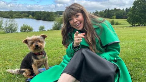 Briony May, wearing a long green coat and sitting on grass beside a lake, pointing at the camera and smiling. She has long dark hair and a fringe. Beside her is a terrier-type dog.