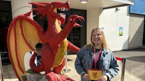 Gaby is standing next to a red and yellow dragon sculpture. She is wearing a grey top and denim jacket and has blonde hair. She is standing in front of Taunton College. 